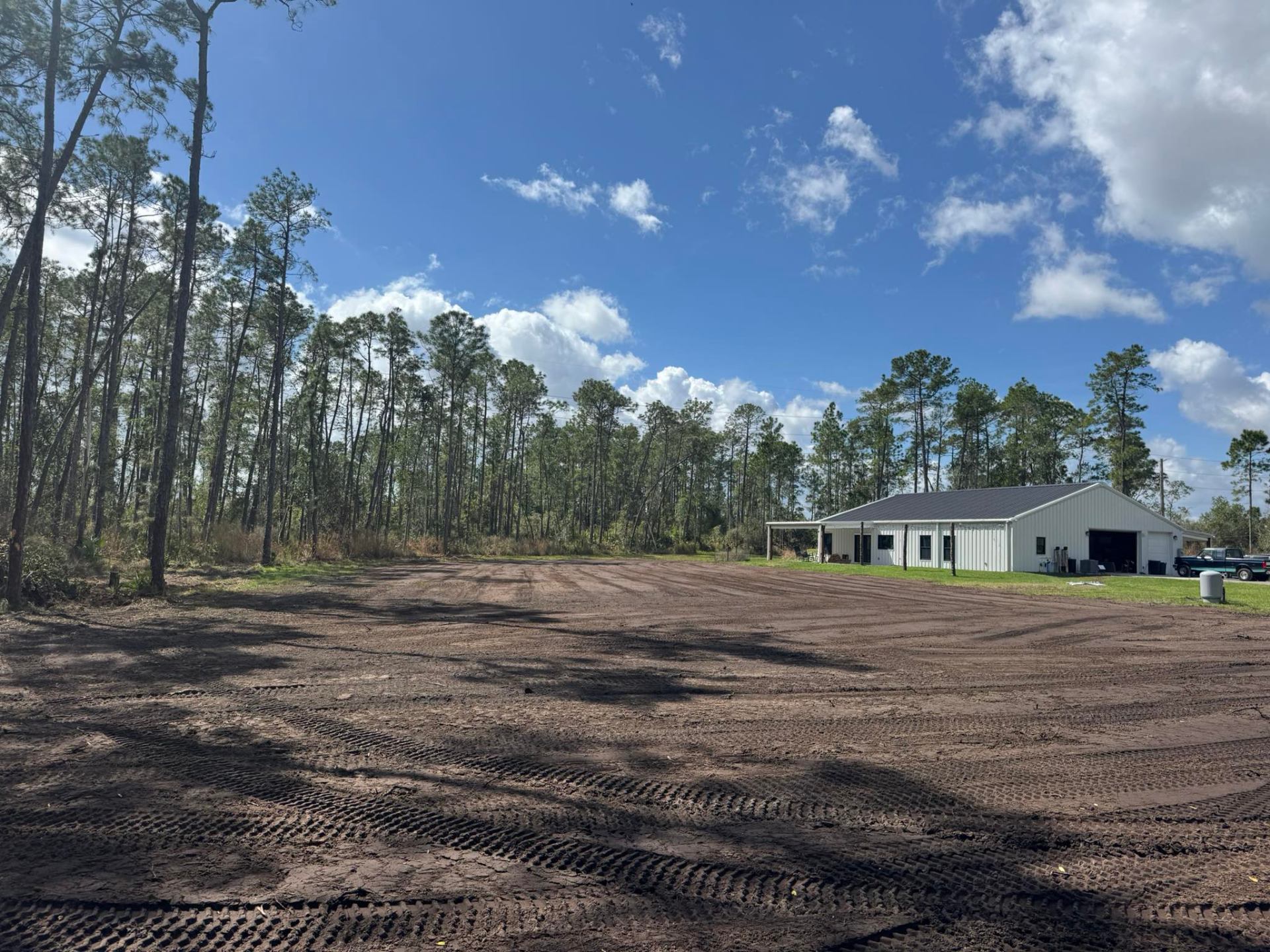 Graded dirt lot with equipment tracks next to white metal building surrounded by pine trees in Florida