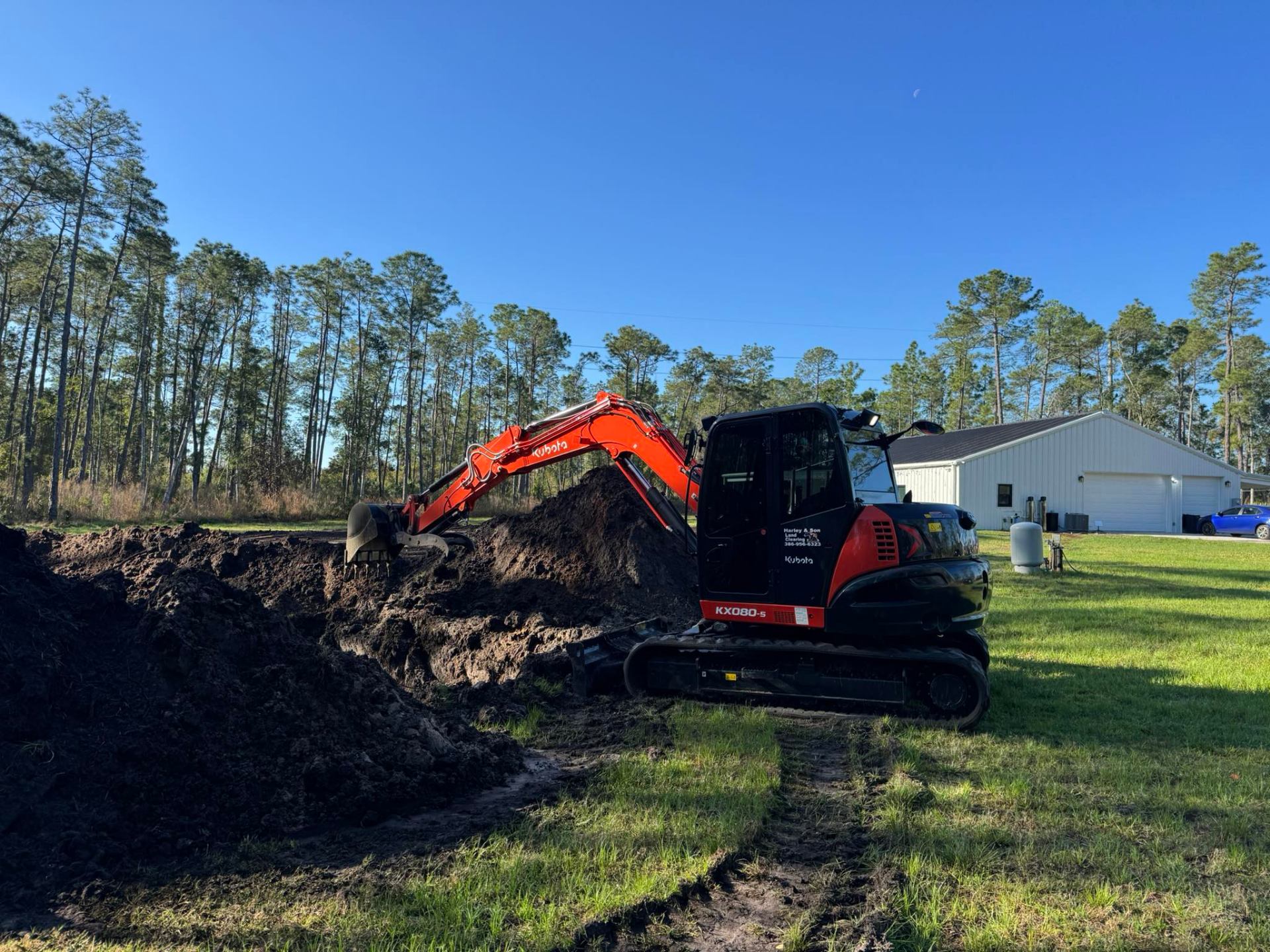 Kubota KX080 excavator digging large dirt pile on green lawn with pine trees in Deltona, FL
